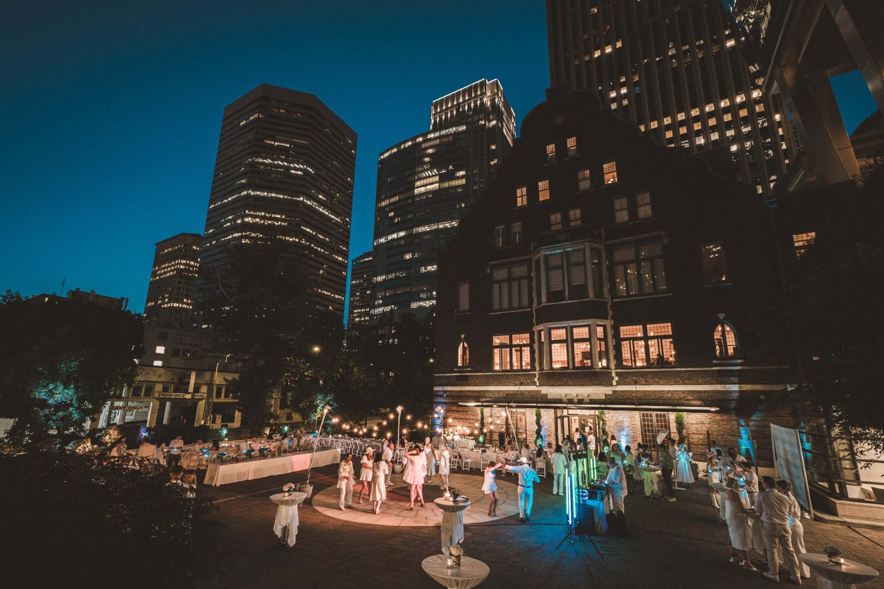Members dressed in white dancing under string lights in the Club's courtyard at dusk, with the clubhouse and downtown towers behind